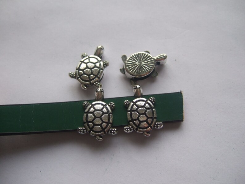 May include: Four silver turtle charms with detailed shell designs. Two turtles are attached to a green leather strap.