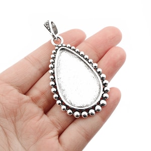 May include: Silver teardrop pendant with a smooth, round bezel for a cameo or other embellishment. The pendant is surrounded by a decorative border of small, round beads.
