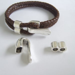 Puede incluir: Un brazalete de cuero marrón con una hebilla plateada y dos cierres plateados. El brazalete está trenzado y tiene un diseño sencillo.