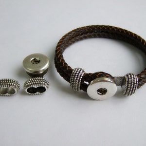 Puede incluir: Un brazalete de cuero marrón con cierre de presión plateado y dos tapas de extremo plateadas. El brazalete está trenzado y tiene un diseño decorativo en las tapas de extremo.