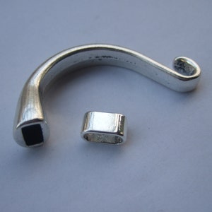 May include: Two silver metal pieces for jewelry making. One piece is a curved, open ring with a square opening. The other piece is a small, rectangular tube.