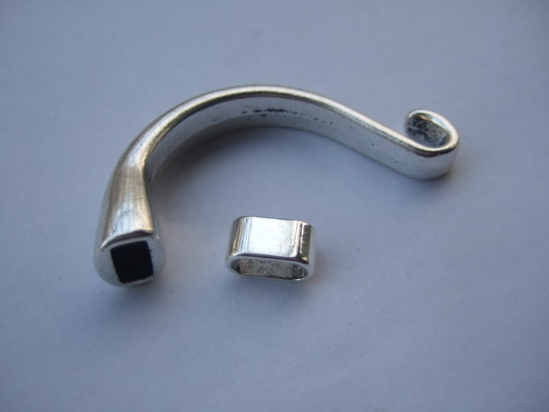 May include: Two silver metal pieces for jewelry making. One piece is a curved, open ring with a square opening. The other piece is a small, rectangular tube.