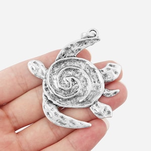 Peut inclure: Pendentif en forme de tortue argenté avec un motif de coquille en spirale. Le pendentif a une finition texturée et une boucle en haut pour l'attacher à une chaîne.