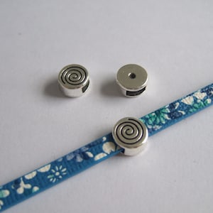 Op de afbeelding: Twee zilveren metalen kralen met een spiraalvormig ontwerp en een zilveren metalen kraal met een gat in het midden. De kralen liggen op een blauw-witte stof met een patroon.