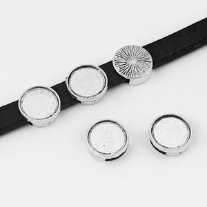 May include: Silver-toned metal round beads with a decorative design. The beads have a flat surface and a groove around the edge. The beads are shown on a black leather strap.