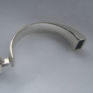 May include: A silver ring with a square-shaped opening on one end and a rounded, flat surface on the other end.