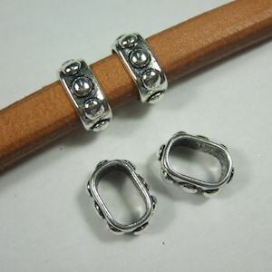 May include: Close-up of three silver-colored metal oval beads with a dotted design. Two beads are on a brown leather cord, and one is loose. The beads have a vintage or antique appearance, suitable for jewelry making or crafts.