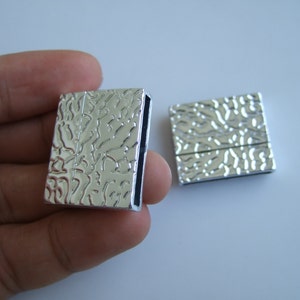 Könnte beinhalten: Zwei silberne Magnetverschlüsse mit einer strukturierten Oberfläche. Die Verschlüsse sind rechteckig und haben eine glatte, polierte Oberfläche.