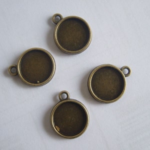 May include: Four antique bronze-coloured round pendant trays with a loop at the top for attaching to a chain or cord. The trays have a recessed area for setting a cabochon or other decorative element. The pendants are arranged on a white surface.