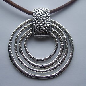 Op de afbeelding: Een zilveren ketting met een hanger in de vorm van een cirkel met meerdere concentrische ringen. De hanger is bevestigd aan een bruin leren koord.
