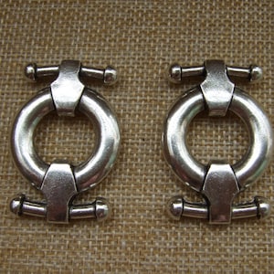 May include: Two silver metal ring clasps with a decorative design. Each clasp has a round ring and a bar with two knobs on either end.