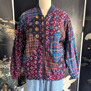 May include: A patchwork bomber jacket with a zipper closure, featuring a mix of patterns in shades of purple, blue, and pink. The jacket is paired with light blue denim jeans. The jacket's design includes floral and geometric shapes.