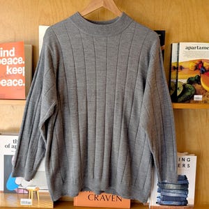 May include: A gray ribbed knit sweater with a mock neck, hanging on a wooden hanger. The sweater is displayed against a backdrop of bookshelves filled with books and magazines.