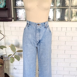 K&ouml;nnte beinhalten: Hellblaue Jeans mit hoher Taille und geradem Bein. Die Jeans befinden sich an einem Mannequin, das einen schwarzen Hut mit goldfarbenem Band tr&auml;gt.