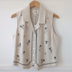 May include: A beige sleeveless vest featuring a notched collar and a front button closure. The garment is decorated with black embroidered floral patterns and a delicate vine-like border along the hem and front edges, hanging on a wooden hanger.