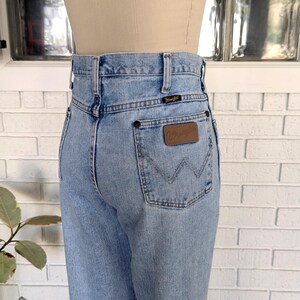 K&ouml;nnte beinhalten: Hellblaue Jeans aus Denim mit hoher Taille und einem braunen Lederpatch mit dem Wort "Wrangler" auf der Ges&auml;&szlig;tasche.