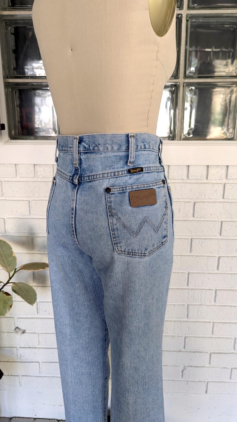 K&ouml;nnte beinhalten: Hellblaue Jeans aus Denim mit hoher Taille und einem braunen Lederpatch mit dem Wort "Wrangler" auf der Ges&auml;&szlig;tasche.