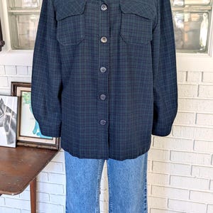 Pode incluir: Uma camisa abotoada xadrez azul escuro e verde com dois bolsos no peito. A camisa é exibida em um manequim e combinada com jeans azul claro. A camisa tem uma gola clássica e mangas compridas.