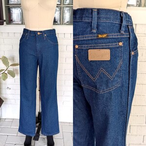 K&ouml;nnte beinhalten: Eine blaue Jeans aus Denim mit einem braunen Leder-Wrangler-Patch auf der Ges&auml;&szlig;tasche. Die Jeans hat einen geraden Schnitt und eine hohe Taille.