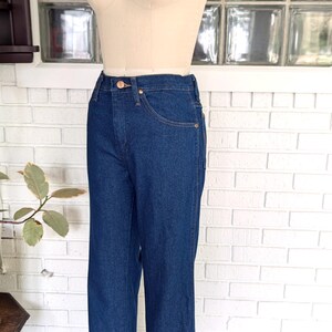 K&ouml;nnte beinhalten: Eine dunkelblaue Jeans mit hoher Taille und geradem Bein. Die Jeans befindet sich an einem Mannequin und wird vor einer wei&szlig;en Backsteinwand pr&auml;sentiert.
