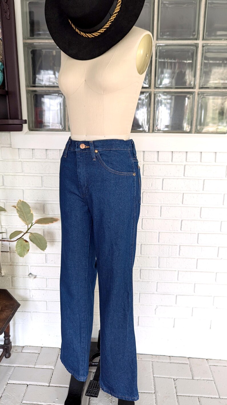 K&ouml;nnte beinhalten: Eine dunkelblaue Jeans mit hoher Taille und geradem Bein. Die Jeans befindet sich an einem Mannequin und wird vor einer wei&szlig;en Backsteinwand pr&auml;sentiert.