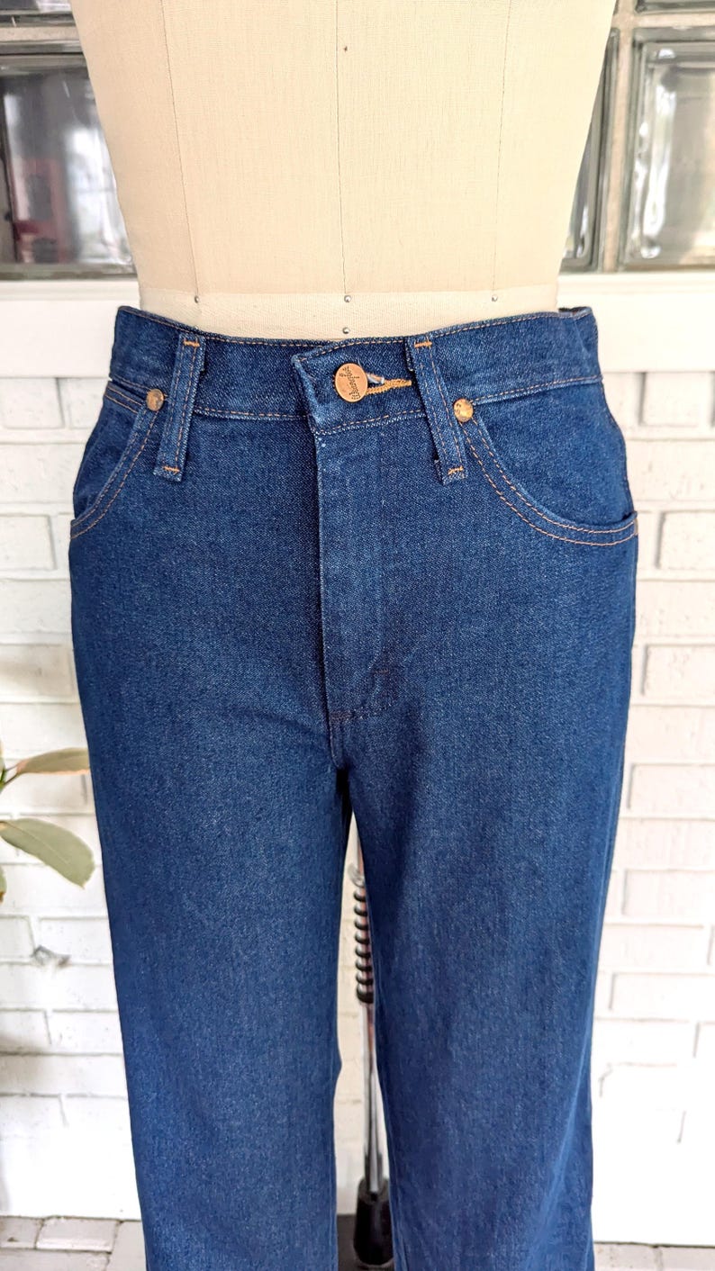 K&ouml;nnte beinhalten: Eine dunkelblaue Jeanshose mit einem braunen Leder-Patch am hinteren Bund, auf dem "Wrangler" steht.