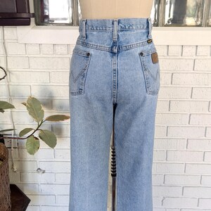 K&ouml;nnte beinhalten: Hellblaue Jeans aus Denim mit hoher Taille und geradem Bein. Die Jeans haben ein braunes Leder-Patch auf der rechten Ges&auml;&szlig;tasche mit dem Wrangler-Logo.