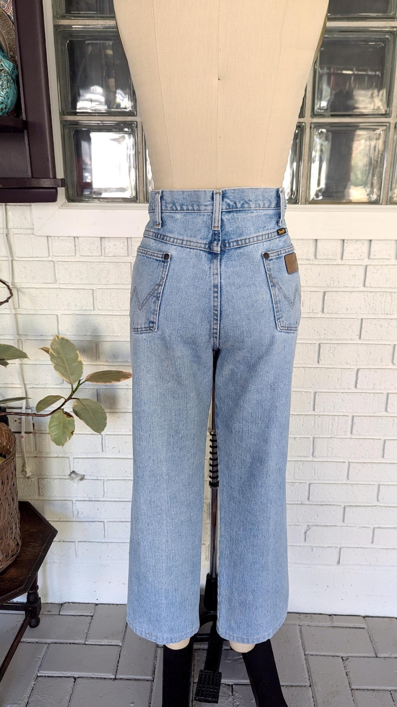 K&ouml;nnte beinhalten: Hellblaue Jeans aus Denim mit hoher Taille und geradem Bein. Die Jeans haben ein braunes Leder-Patch auf der rechten Ges&auml;&szlig;tasche mit dem Wrangler-Logo.