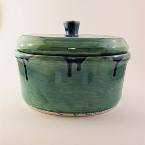 May include: A green ceramic lidded jar with a white rim and blue glaze accents. The jar has a smooth, glossy finish and a rounded shape.