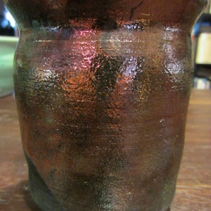 May include: A small, dark brown ceramic jar with a textured surface and iridescent glaze. The jar has a rounded body and a wide mouth.