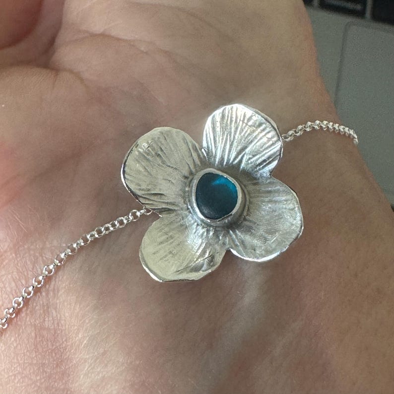 May include: A silver flower bracelet with a blue gemstone centre. The flower has textured petals and is attached to a delicate chain. The bracelet is a piece of jewellery.