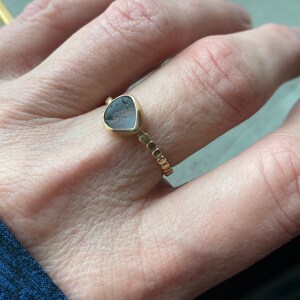 May include: A gold ring featuring a unique, irregularly shaped, gray-blue stone. The stone is set in a gold bezel, and the band has a delicate beaded design. The ring is shown on a person's finger, highlighting its elegant and artistic design.