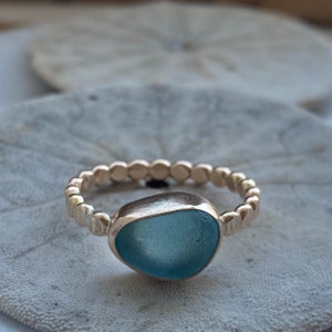 May include: A gold-colored ring featuring a light blue, irregularly shaped stone. The band is adorned with small, evenly spaced circular details. The ring is set against a neutral, textured background, highlighting the jewelry's design.