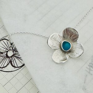 May include: A silver flower pendant necklace with a turquoise centre stone. The four-petal flower has textured details and hangs from a delicate chain. The necklace is displayed on a white surface.