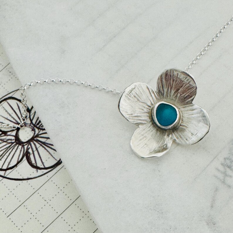 May include: A silver flower pendant necklace with a turquoise centre stone. The four-petal flower has textured details and hangs from a delicate chain. The necklace is displayed on a white surface.