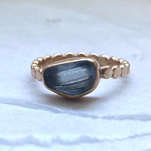 May include: A gold ring featuring a large, irregularly shaped, blue-gray gemstone. The band has a textured, beaded design. The gemstone is set in a gold bezel, and the ring is displayed on a white surface.