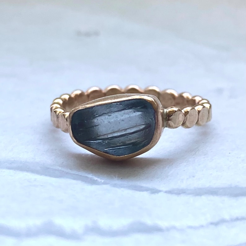 May include: A gold ring featuring a large, irregularly shaped, blue-gray gemstone. The band has a textured, beaded design. The gemstone is set in a gold bezel, and the ring is displayed on a white surface.
