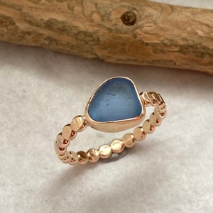 May include: A rose gold ring featuring a light blue, sea glass-like stone. The band is adorned with small, circular accents. The ring is set against a neutral background, highlighting the jewelry's design.