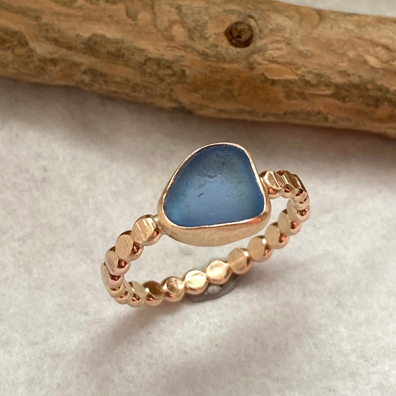 May include: A rose gold ring featuring a light blue, sea glass-like stone. The band is adorned with small, circular accents. The ring is set against a neutral background, highlighting the jewelry's design.