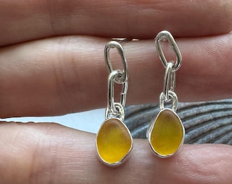 May include: A pair of silver dangle earrings with yellow sea glass drops. The earrings feature a silver chain with a small loop at the top.