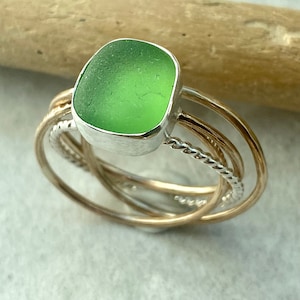May include: A silver ring with a green sea glass stone set in the centre. The ring is surrounded by three gold bands, two plain and one twisted.