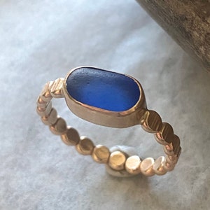 May include: A gold-colored ring featuring a rectangular, deep blue sea glass stone. The band has a textured, beaded design. The ring is set against a neutral background, highlighting the contrast between the metal and the glass.