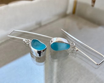 May include: A pair of silver dangle earrings featuring light blue sea glass. The earrings are made with a simple silver wire and a small silver bead at the end.