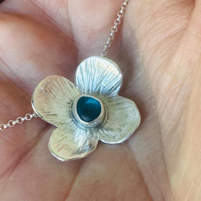May include: A silver flower pendant necklace with a textured finish. The pendant has a turquoise-coloured stone at its centre. The necklace is on a delicate silver chain, suitable for everyday wear.