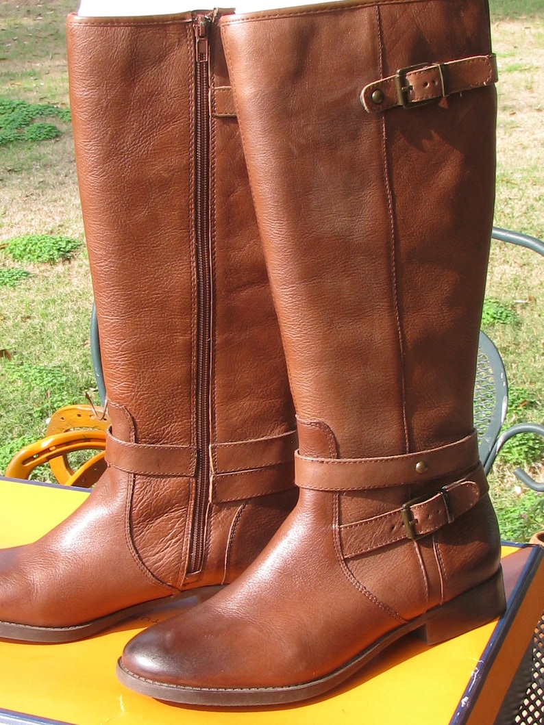 May include: A pair of brown leather knee-high boots with buckles and zippers. The boots have a low heel and a rounded toe.