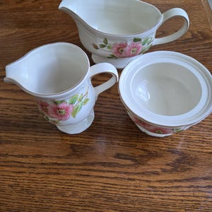 May include: A cream-colored porcelain serving set featuring a gravy boat, creamer, and sugar bowl. Each piece is adorned with a floral pattern of pink flowers and green leaves, with a gold trim. The set is displayed on a wooden surface.