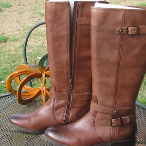 May include: A pair of brown leather riding boots with a buckle strap and zipper closure. The boots have a low heel and are made of a soft, supple leather.