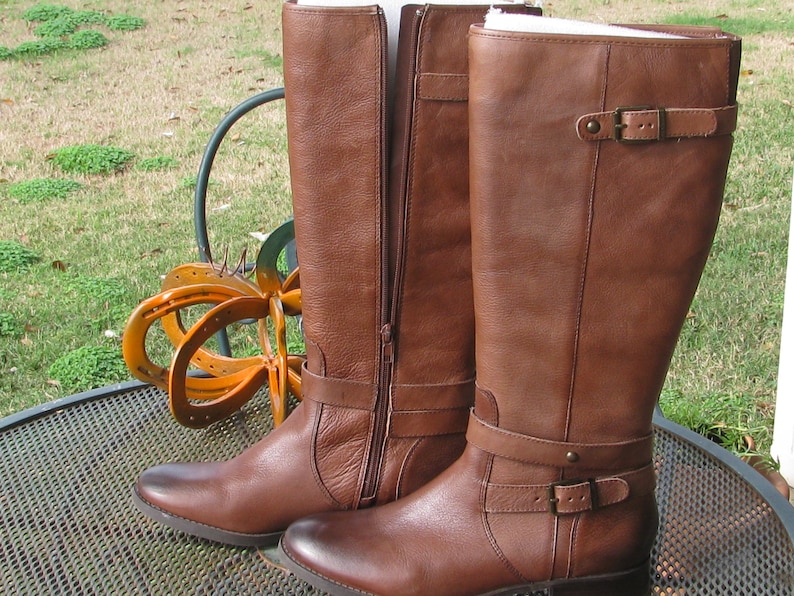 May include: A pair of brown leather riding boots with a buckle strap and zipper closure. The boots have a low heel and are made of a soft, supple leather.