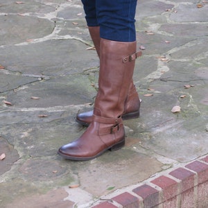 May include: Brown leather riding boots with a buckle detail. The boots are worn on a stone pathway with fallen leaves.