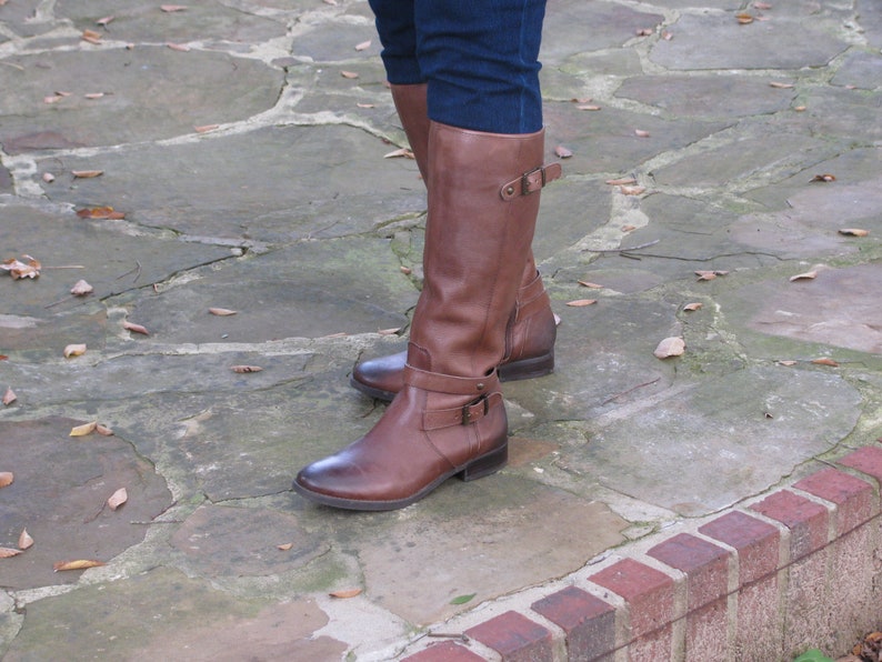 May include: Brown leather riding boots with a buckle detail. The boots are worn on a stone pathway with fallen leaves.